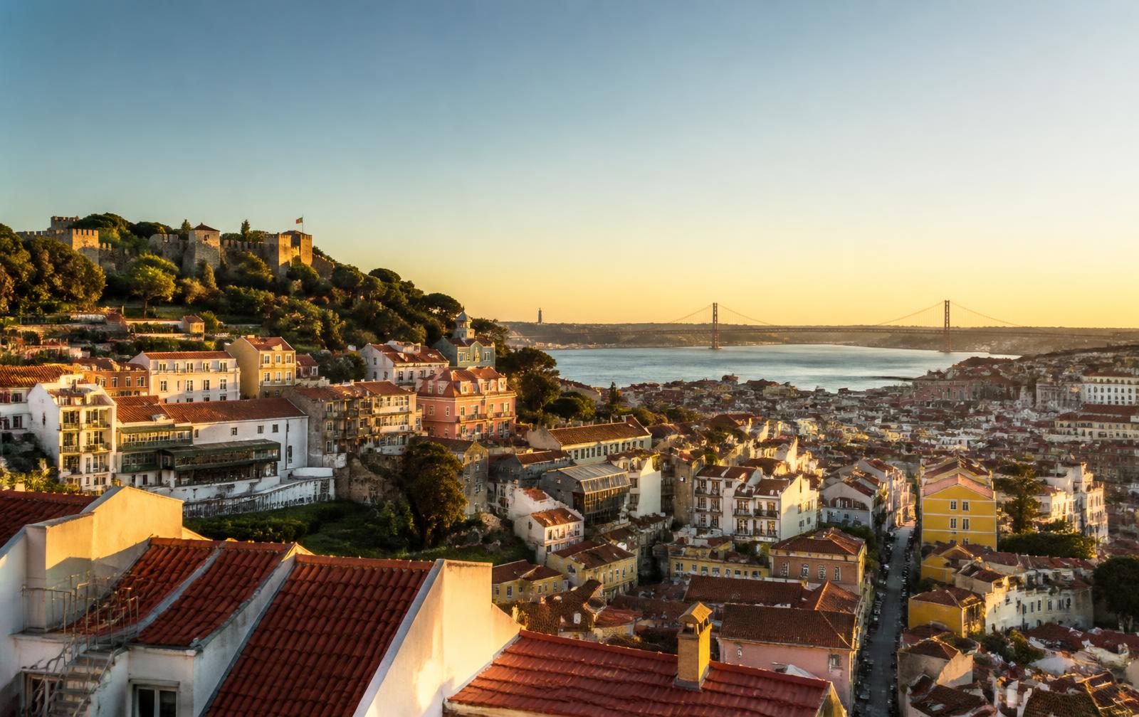 Lisbon's red rooftops and hills rolling down to the river in golden evening light, seen from above